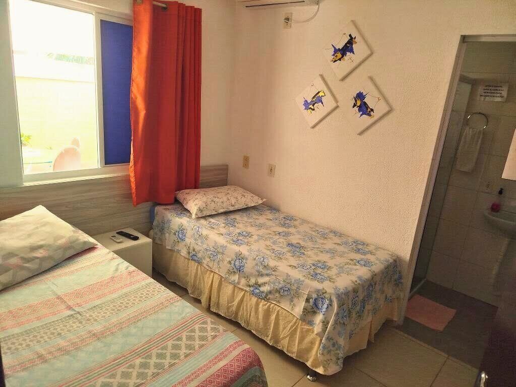 Ein Schlafzimmer mit einem Bett, einem zusätzlichen Bett, einem Fenster mit Vorhängen, einem an der Wand befestigten Kunstwerk und einer Badezimmertür.