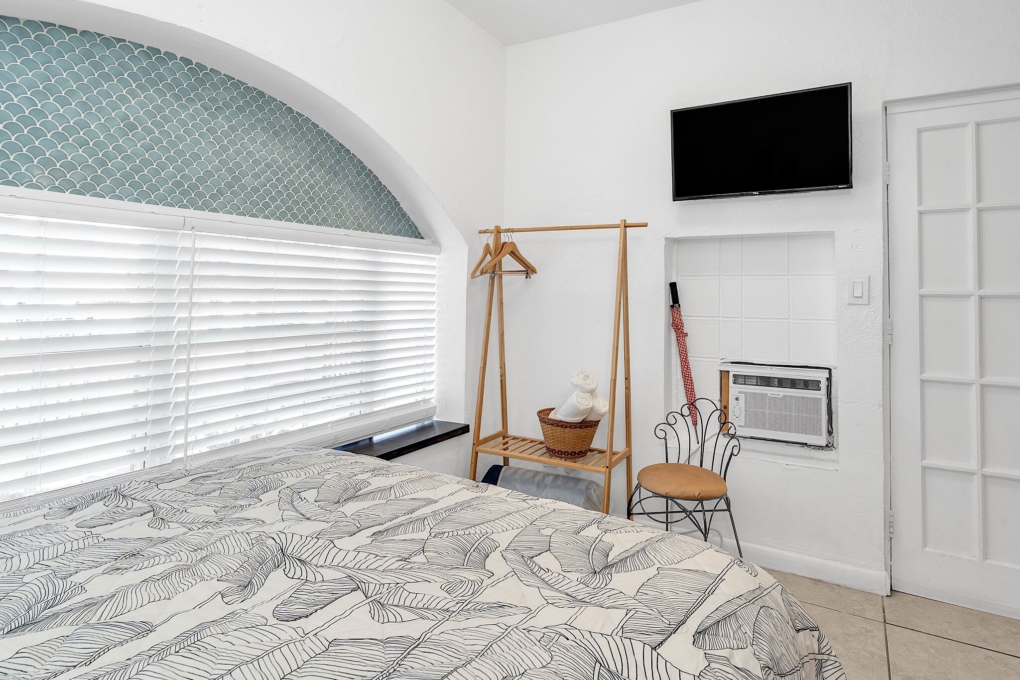 A bedroom with a bed, a television, a window with blinds, a wall-mounted air conditioning unit, and a wooden hanger stand with clothes.