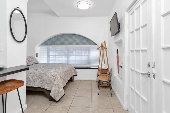 A bedroom with a bed, a chair, a TV, a window with blinds, and a door.