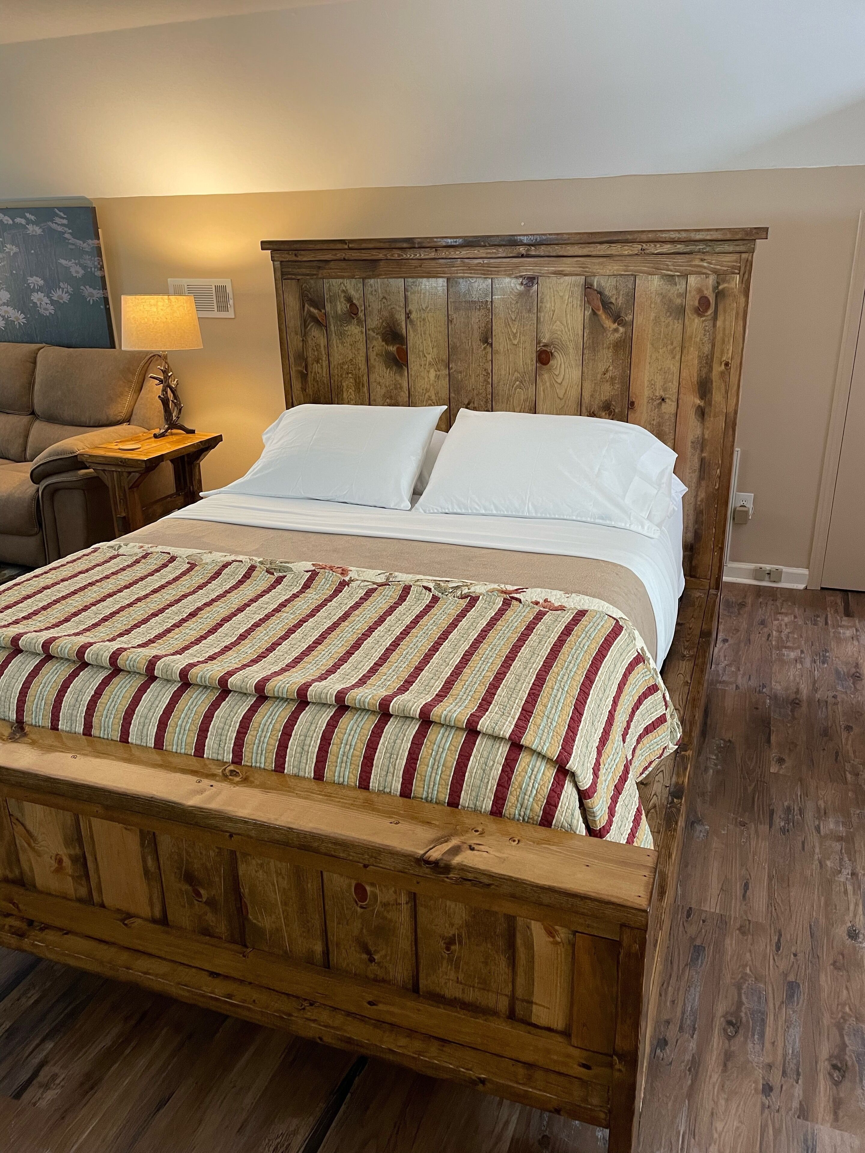 Ein Bett mit Holzbettgestell, weißer Bettwäsche und einer Streifen-Decke, ein Nachttisch mit Lampe sowie ein Holzboden.