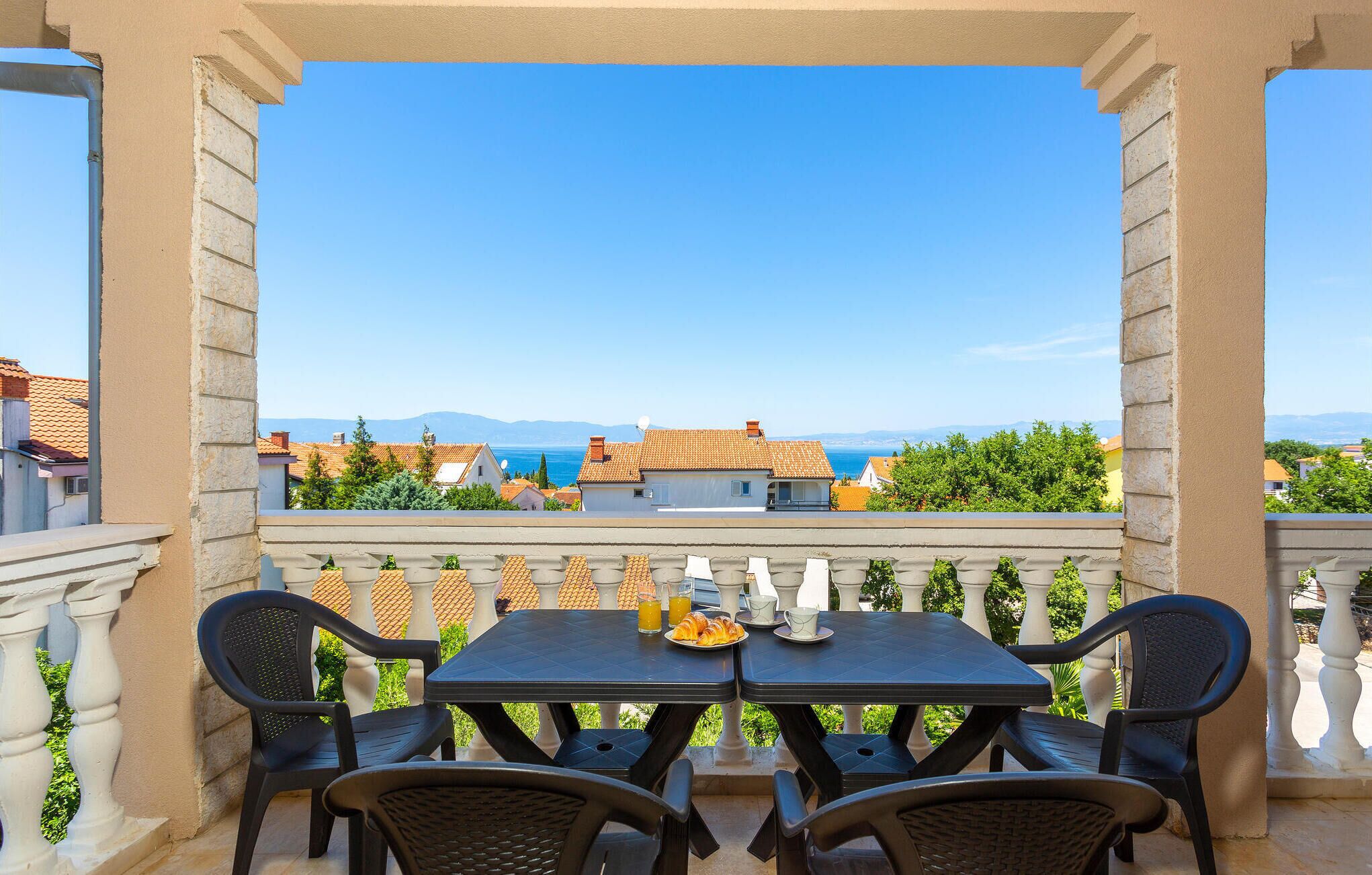 Ein Balkon mit Tisch und Stühlen, mit Blick auf eine Küstenstadt mit Häusern und blauem Himmel.