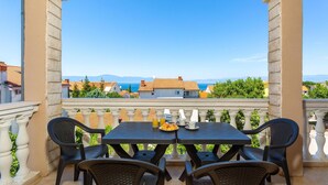 A balcony with a table and chairs, overlooking a coastal town with houses and a clear blue sky. - Cozy apartment in Malinska with WiFi (Malinska)