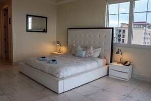 A bedroom with a large bed, two bedside tables, a nightstand, a mirror, and a window with a city view.