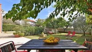 Une table en bois sur laquelle repose un panier de fruits, entourée de chaises rouges, dans un jardin avec une pelouse et un mur de briques en arrière-plan.