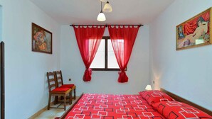 Une chambre à coucher avec un lit, une chaise, une fenêtre avec des rideaux rouges et deux lampes fixées au mur.