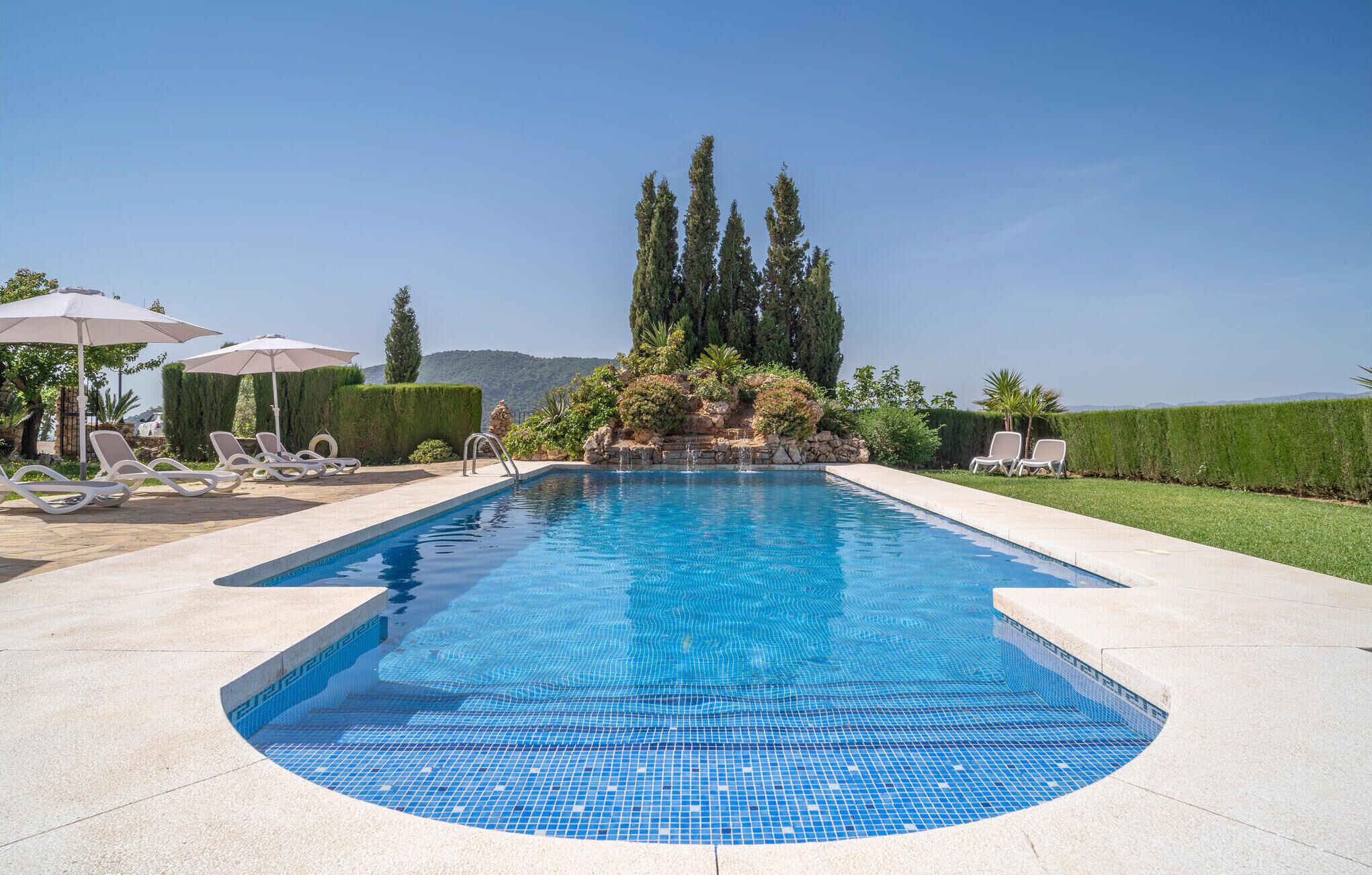 Une piscine rectangulaire bordée de carreaux, entourée de chaises longues et de parasols, avec un jardin aménagé et des arbres en arrière-plan.