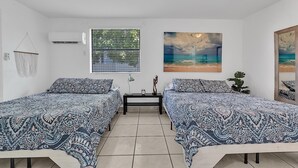 A bedroom with two beds, a window with blinds, an air conditioner, and a painting of a beach scene. - Little Havana Studio for 4 • Free Parking (Miami)