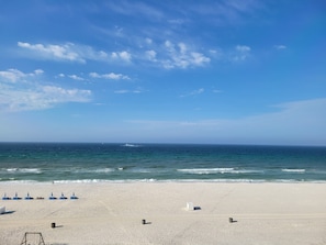 Uma praia com céu claro, mar calmo e algumas pessoas caminhando.