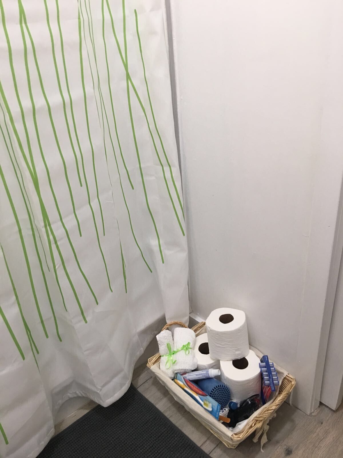 Un baño con cortina de ducha blanca con líneas verticales verdes, una cesta de mimbre con artículos de aseo y un rollo de papel higiénico.
