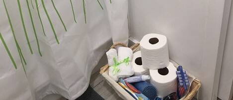 Un baño con cortina de ducha blanca con líneas verticales verdes, una cesta de mimbre con artículos de aseo y un rollo de papel higiénico.