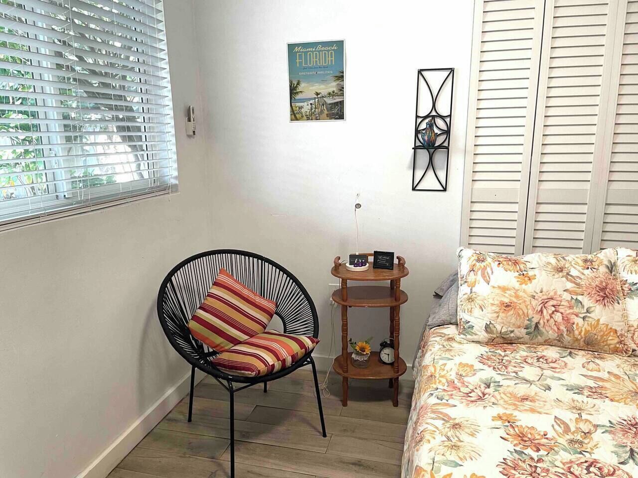 Una habitación con cama, una silla, una mesita de madera, un reloj de pared y un cartel.