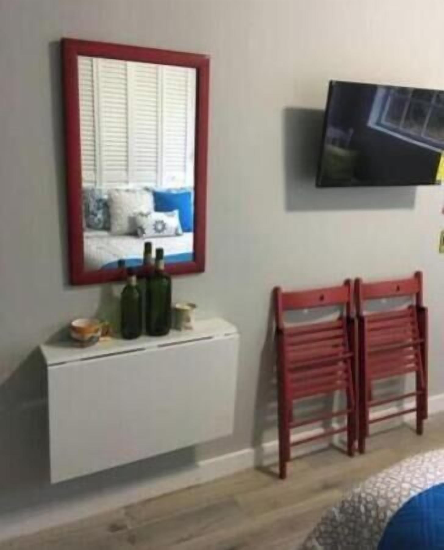 A room with a white cabinet, a red-framed mirror, a television on the wall, and a pair of red chairs.