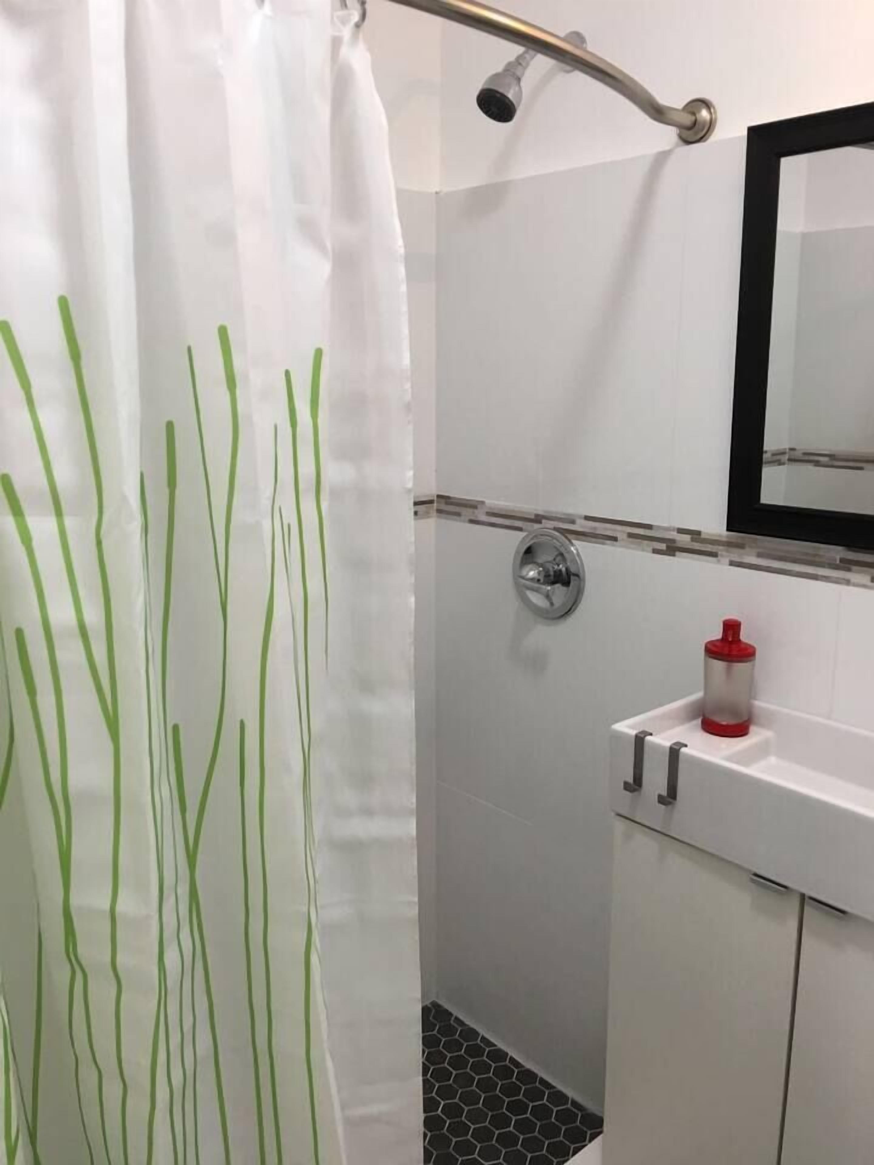 Área de duche com parede revestida a azulejos brancos, um chuveiro, um separador de duche de vidro com linhas verdes, um dispensador de sabonete vermelho e branco, e um espelho sobre a pia.