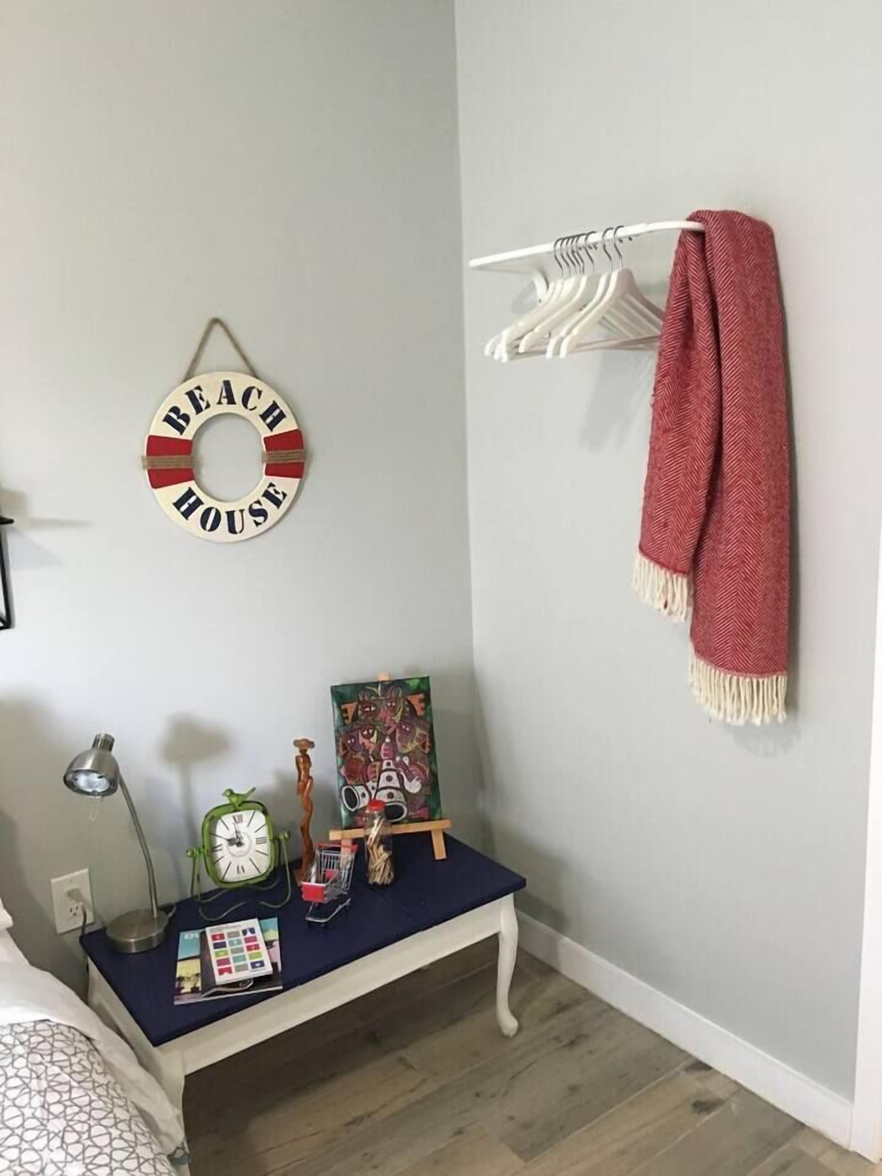 Um quarto com um cabide fixado na parede, um banco com relógio e objetos decorativos, e um letreiro com a inscrição 'BEACH HOUSE'.