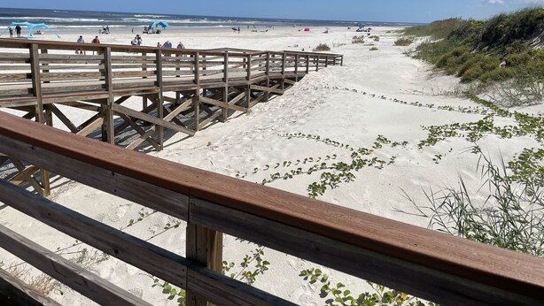 Une promenade en bois mène à une plage de sable où la végétation est clairsemée.