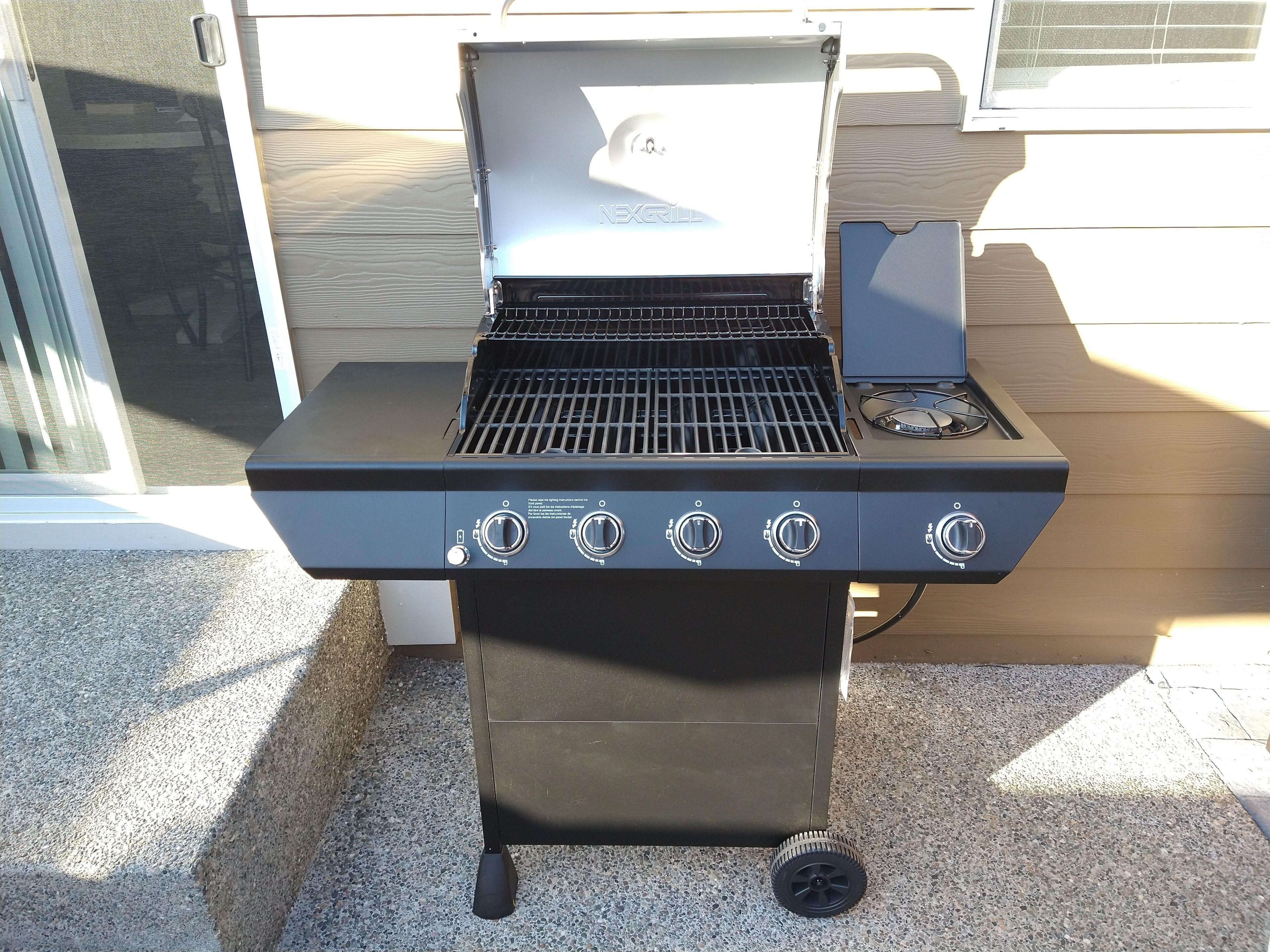 Een draagbare grill met vier bedieningsknoppen en een extra brander, geplaatst op een betonnen terras.