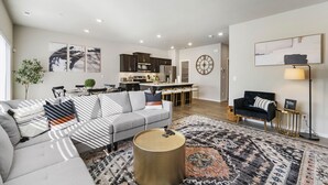 Un salon moderne avec un canapé gris, une table basse ronde et un tapis à motifs. On y trouve également un coin repas avec une table et des chaises, et une cuisine en arrière-plan.