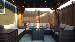Une terrasse extérieure avec des meubles en osier, une table dotée d’un foyer et des guirlandes lumineuses.
