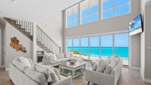 A spacious living room with a large window overlooking a beach, a staircase with a wooden finish, and a flat-screen TV mounted on the wall. - Shades of Blue a luxury home on private beachfront sleeps 20ppl, chef, boat tour (Forbes Hill)