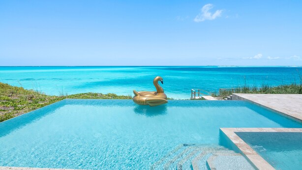 A clear blue pool with a swan float, surrounded by a stone deck and a view of the ocean. - Shades of Blue a luxury home on private beachfront sleeps 20ppl, chef, boat tour (Forbes Hill)