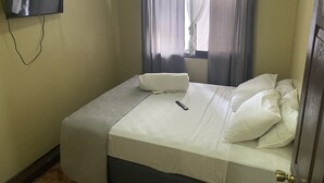 Un petit lit bien fait, avec du linge de lit blanc et des oreillers, une télécommande posée sur le lit et un téléviseur fixé au mur.