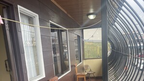 Un espace extérieur abrité, doté d’un banc et de chaises en bois, d’un sol carrelé et offrant une vue sur la rue à travers une rambarde métallique incurvée.