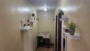 Un couloir étroit aux murs de couleur beige, doté d’un sol carrelé et de quelques étagères blanches où sont disposées des plantes en pot et de petits objets.