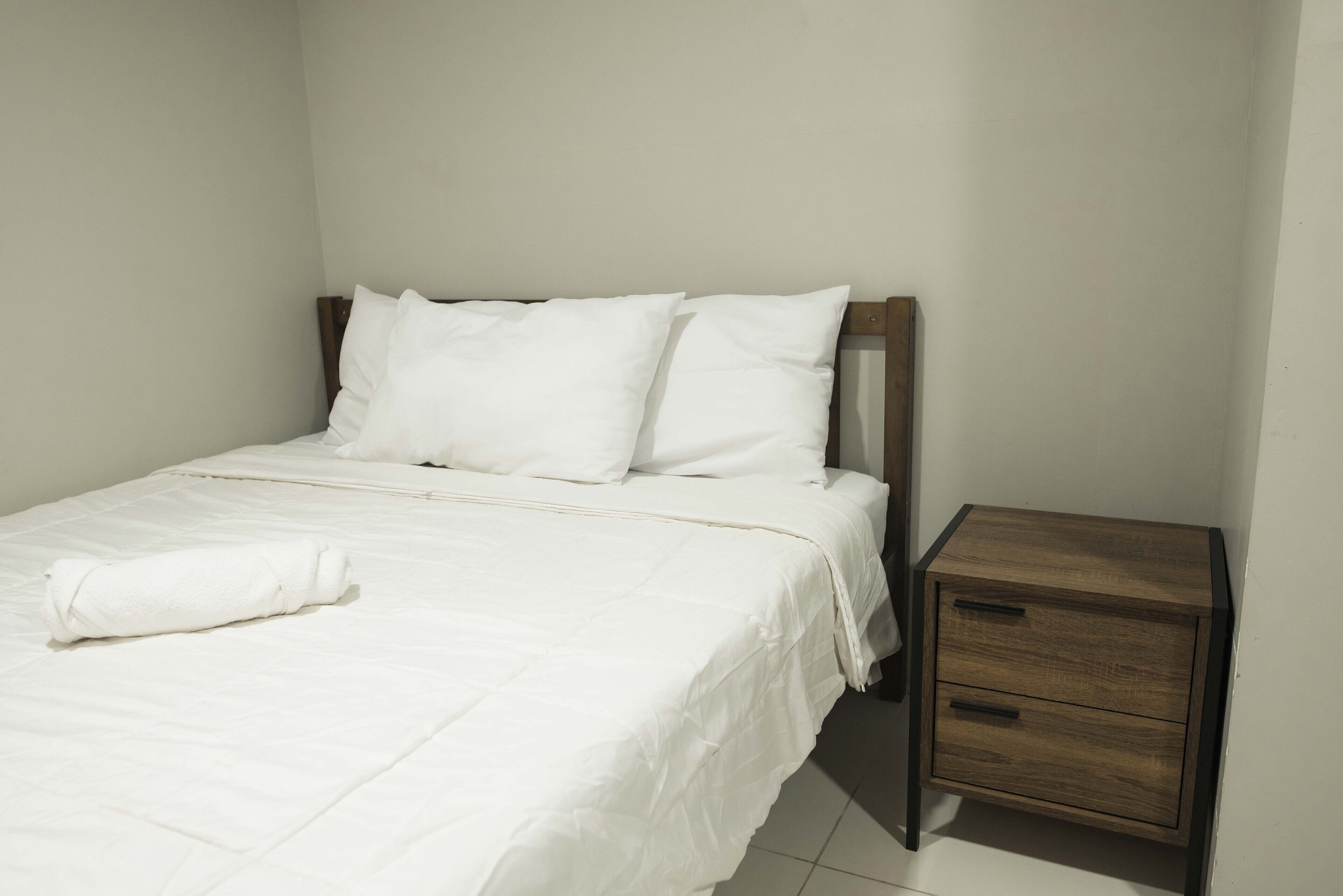 A neatly made bed with white bedding and pillows, a wooden nightstand with drawers, and a plain white wall.
