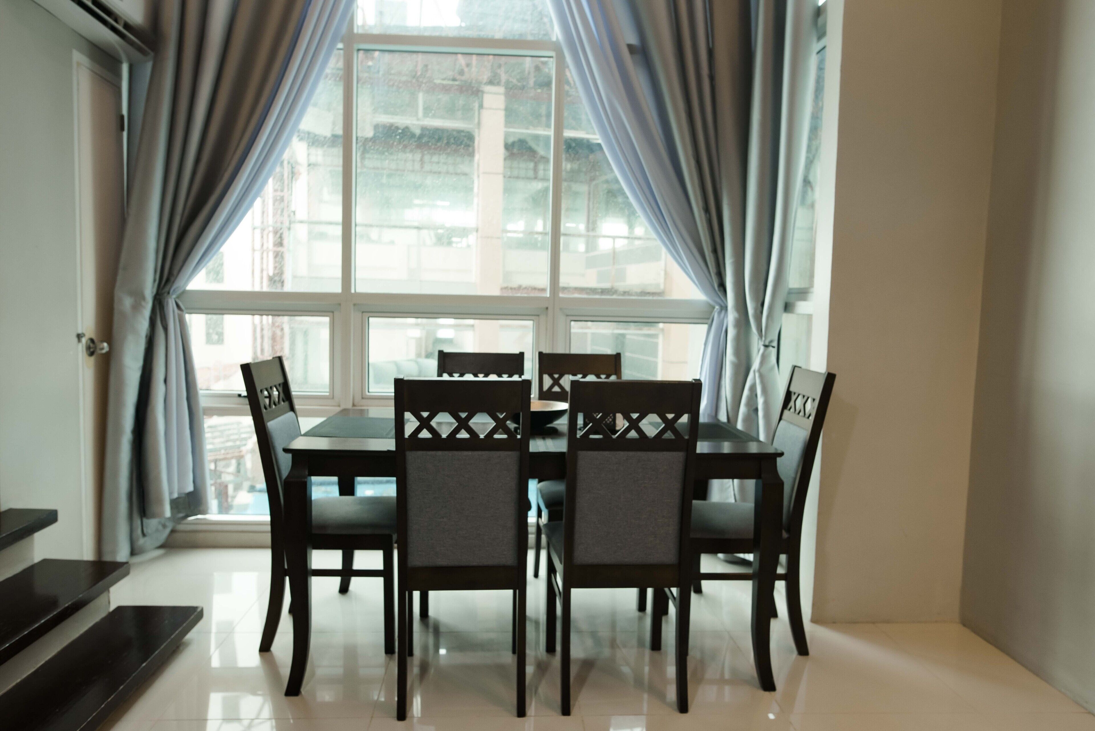 A dining area with a table and chairs, large windows with curtains, and a view of buildings outside.
