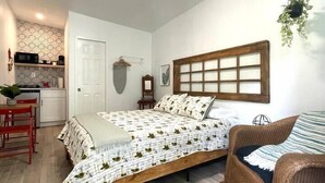 Een slaapkamer met een bed, een stoel, een hangende plant, een hoofdeinde en een keuken op de achtergrond.