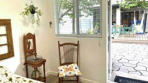 Een kamer met een houten stoel, een hangplant, een raam met zonwering en uitzicht op een terras.