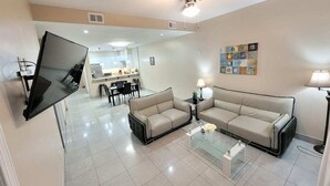 A modern living room with a flat-screen TV mounted on the wall, two beige sofas, a glass coffee table, and a ceiling fan. - Jellyfish 2 BR Oceanfront Condo with Pool (Myrtle Beach)