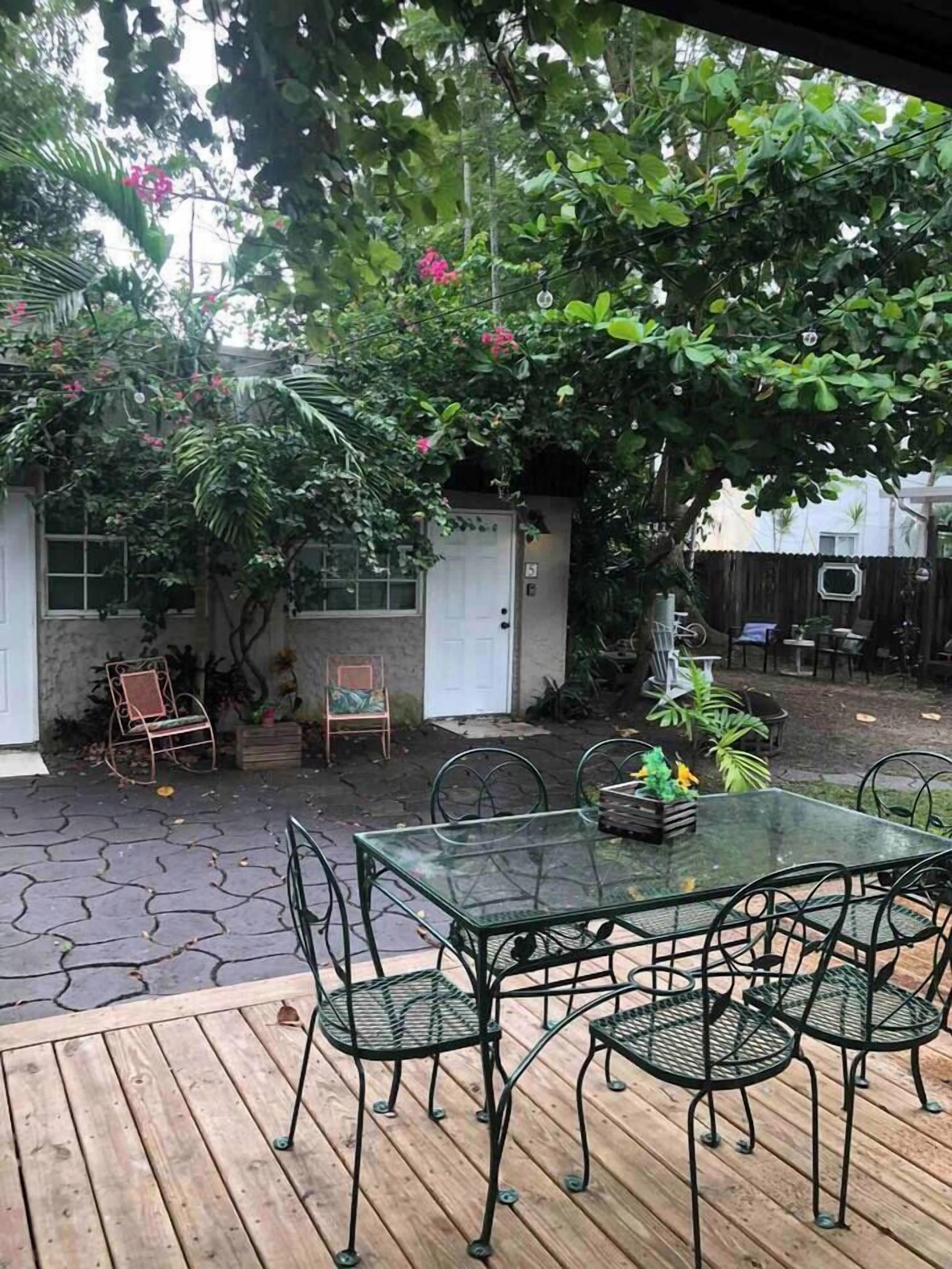 A patio with a glass table and chairs, surrounded by greenery and a wooden deck.