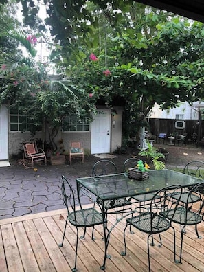 A patio with a glass table and chairs, surrounded by greenery and a wooden deck.