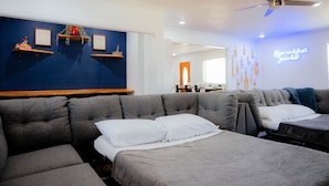 Una moderna camera d'albergo con un ampio divano angolare grigio, un letto con lenzuola bianche e una ventola a soffitto.