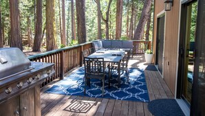 An outdoor deck with a grill, dining table, and chairs, surrounded by tall trees. - Family Retreat! Hot Tub, King Beds & EV Charger (Arnold)