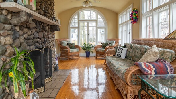 A cozy living room with a stone wall, wicker furniture, a fireplace, and large windows.