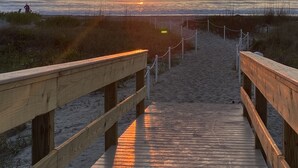 Ein hölzerner Steg führt zu einem Strand, im Hintergrund ist ein Sonnenuntergang zu sehen.