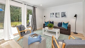 Ein modernes Wohnzimmer mit einer Couch, Sesseln, einem Couchtisch und einem Bücherregal. Große Fenster mit Vorhängen bieten einen Blick ins Freie, und eine Stehlampe sorgt für Licht.