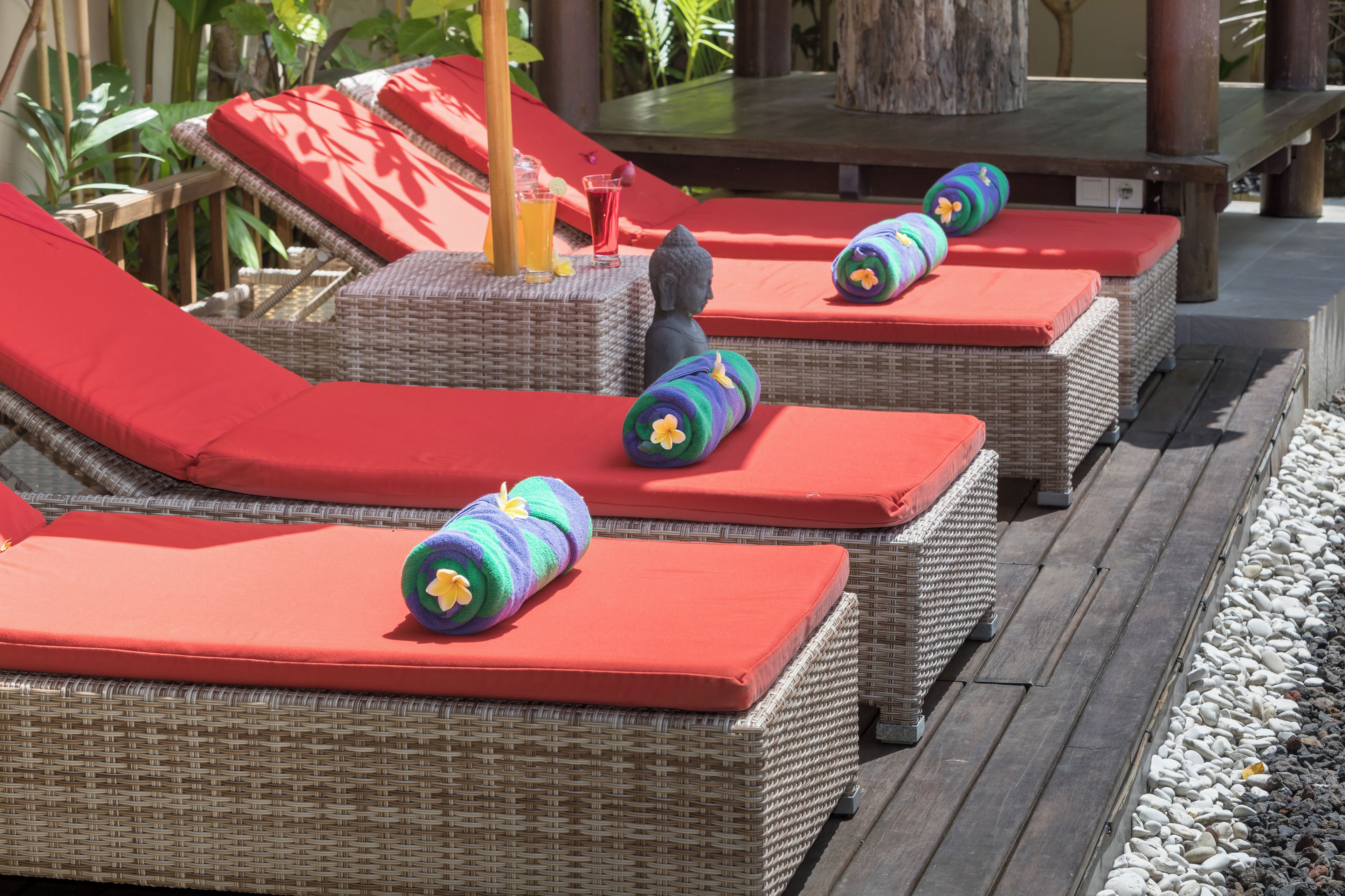 Wicker lounge chairs with red cushions and colorful towels, arranged on a wooden deck with a tropical backdrop.