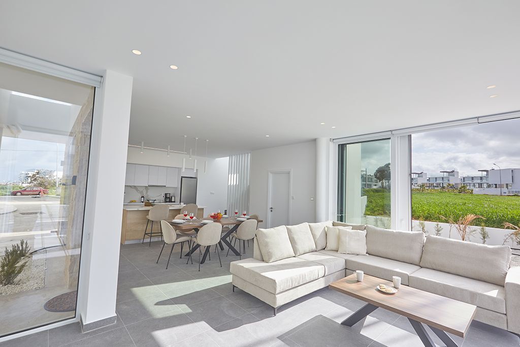 A modern living room with a sectional sofa, a dining area with a table and chairs, and large windows offering a view of a green landscape.