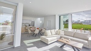 Una sala de estar moderna con un sofá modular, un comedor con mesa y sillas, y grandes ventanales que ofrecen vistas a un paisaje verde.