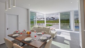 Un comedor moderno con una mesa grande dispuesta para seis comensales, ventanas amplias que ofrecen vistas a una zona residencial y un rincón de estar acogedor con sofá y una mesita.
