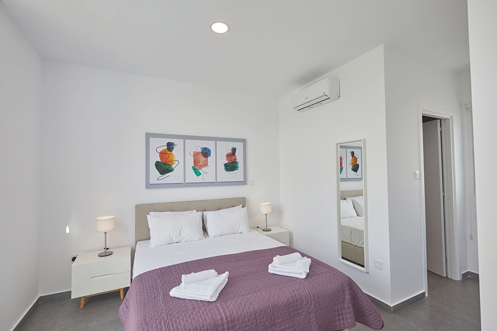 A modern hotel room with a large bed, white walls, and a ceiling light. There are two bedside tables with lamps and a large mirror on the wall.