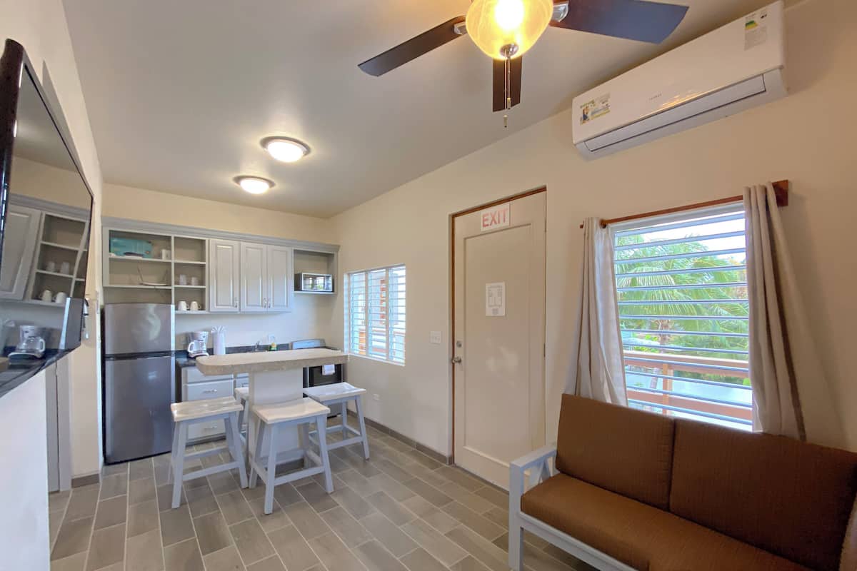 Cocina compacta con refrigerador, lavamanos y gabinetes blancos. Hay un comedor con mesa y sillas, y un sofá marrón.
