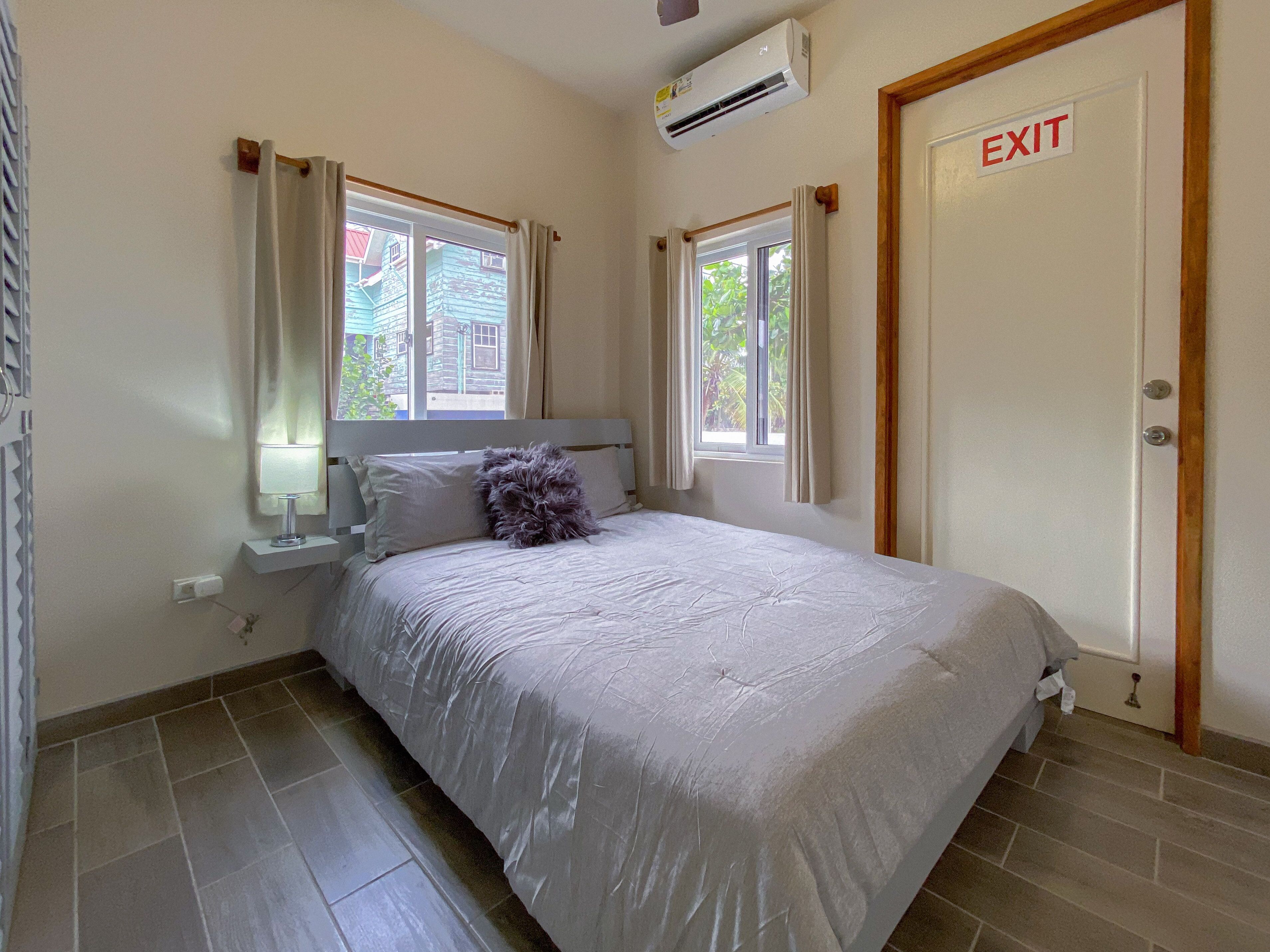 A bedroom with a bed, an air conditioning unit, a window with curtains, and an exit door.