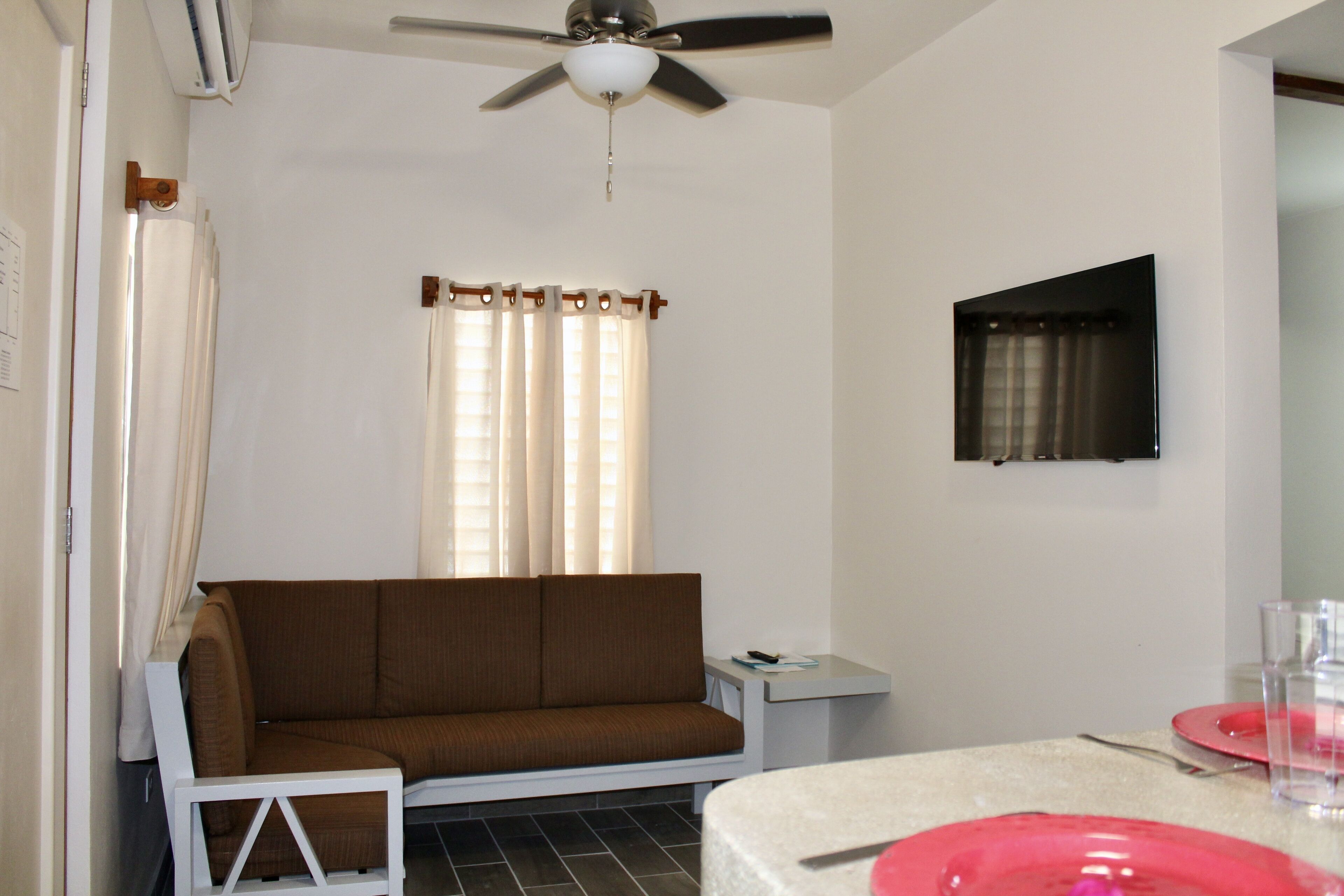 Une pièce avec un canapé marron, une petite table blanche, un ventilateur de plafond et un téléviseur fixé au mur.