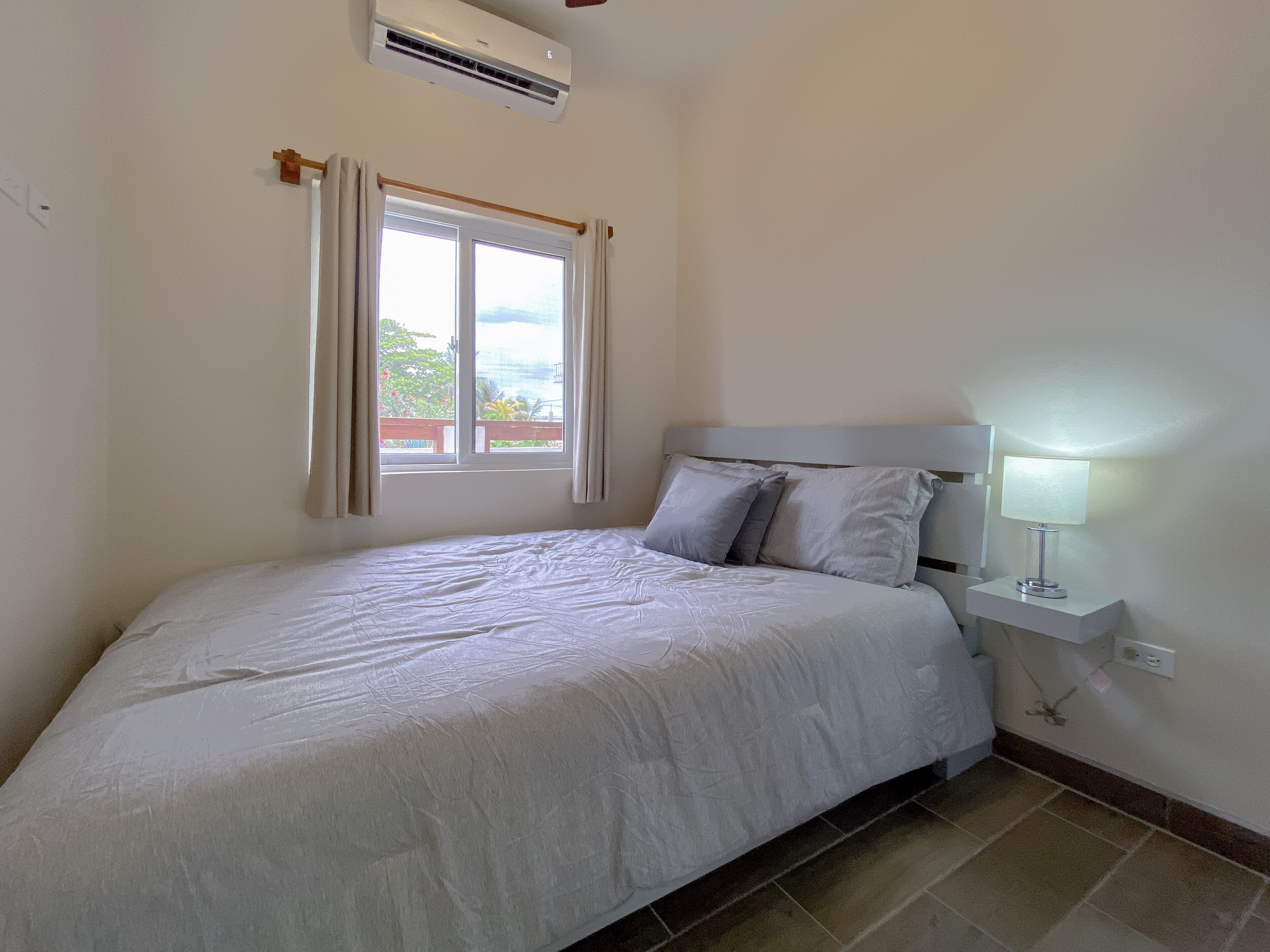 A neatly made bed with a white comforter and pillows, a bedside lamp, a window with curtains, and an air conditioning unit on the wall.