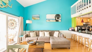 Una sala de estar con pared verde azulado, un sofá seccional gris, una mesa de centro de madera y una cocina con encimera blanca y taburetes de madera.