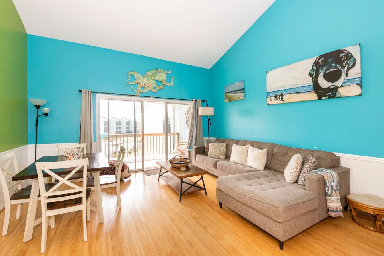 A living room with a blue wall, a grey sofa, a dining table with chairs, and a large window.
